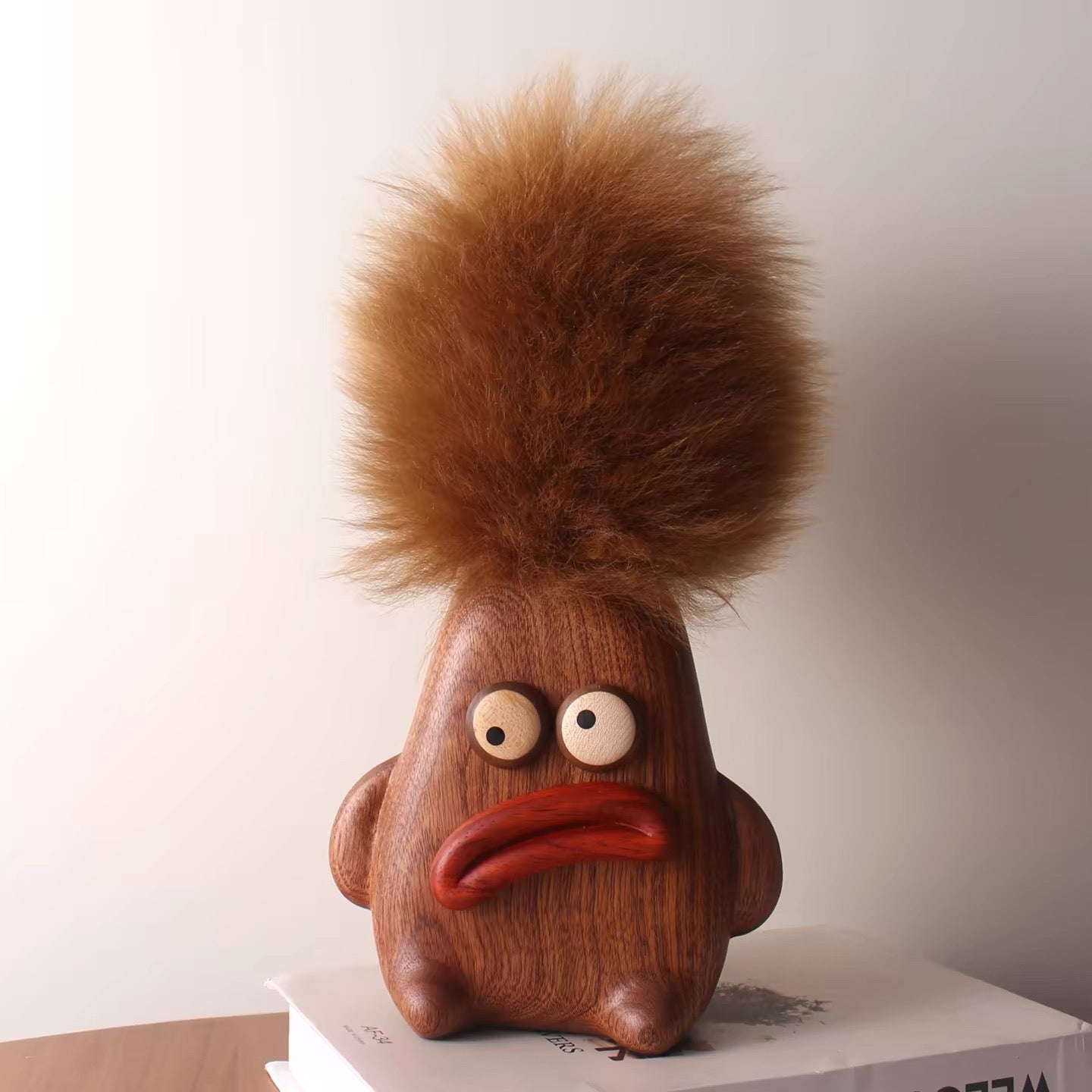 Pouty wooden character holder with a fluffy brown lambswool duster displayed on stacked books.