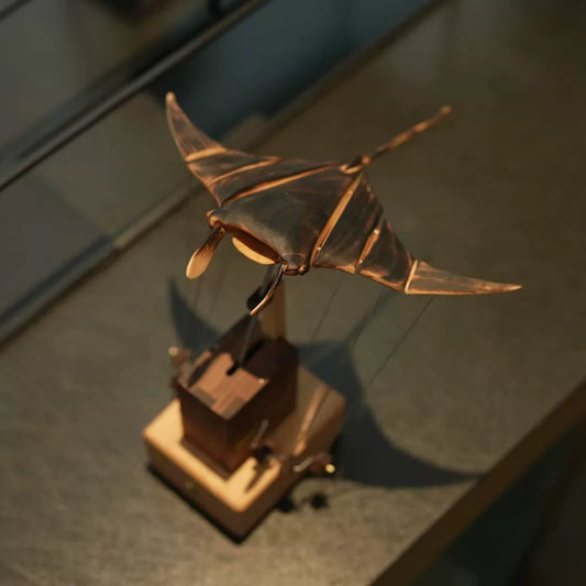 Angled view of a wooden manta ray automaton showing its articulated wings, long tail, and mechanical base.