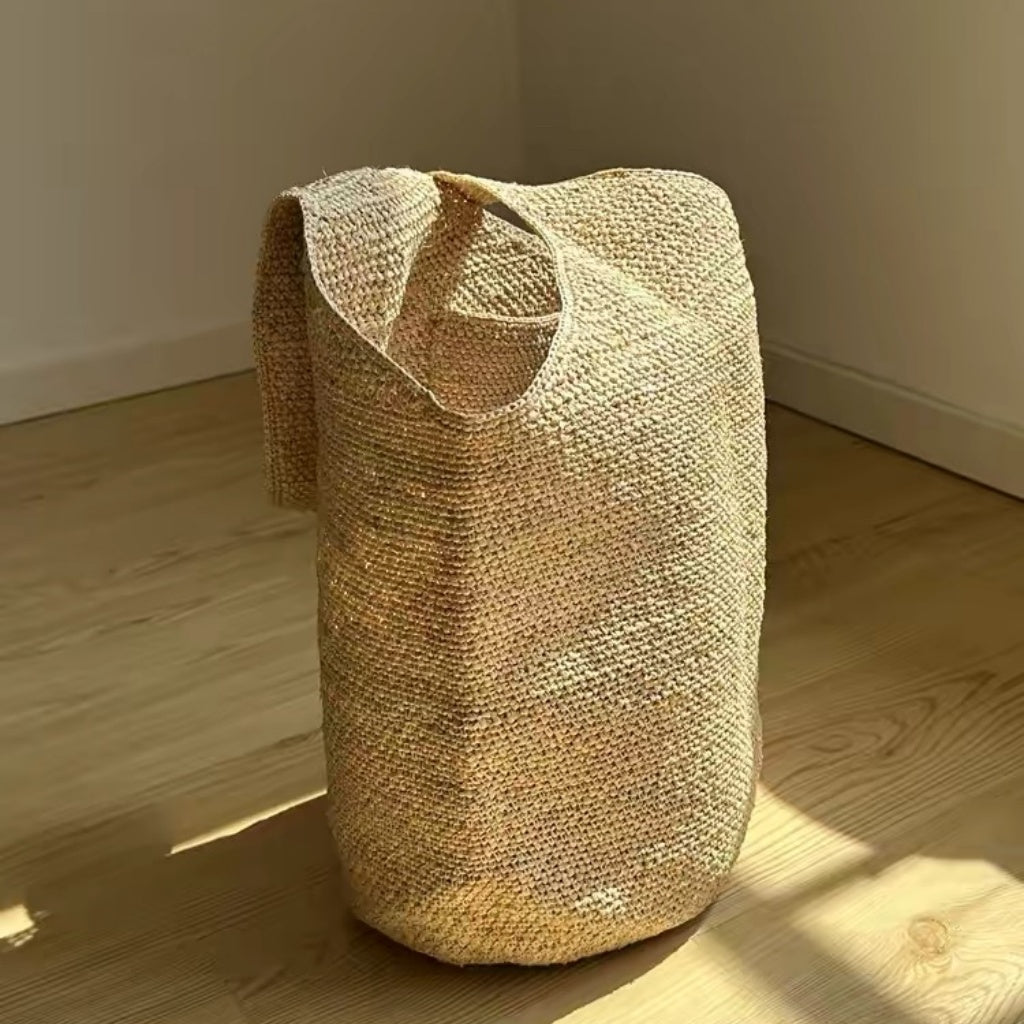 Handwoven natural raffia storage basket with soft handles, photographed in warm sunlight on a wooden floor, perfect for home decor, laundry, blankets, toys, or everyday storage