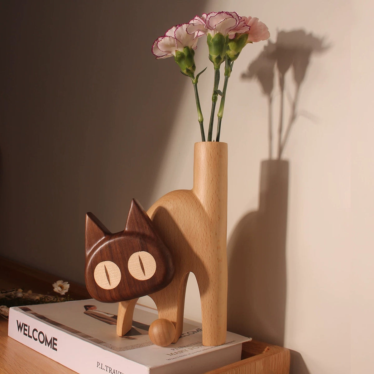 Light beech wooden cat vase with a dark contrasting head and pink flowers on a shelf.