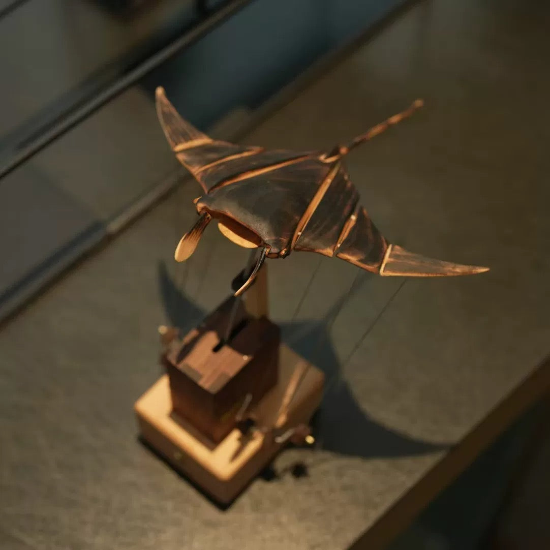 Angled view of a wooden manta ray automaton showing its articulated wings, long tail, and mechanical base.