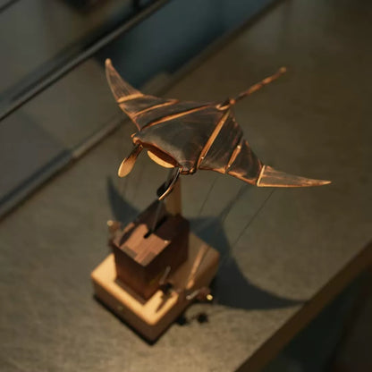 Angled view of a wooden manta ray automaton showing its articulated wings, long tail, and mechanical base.