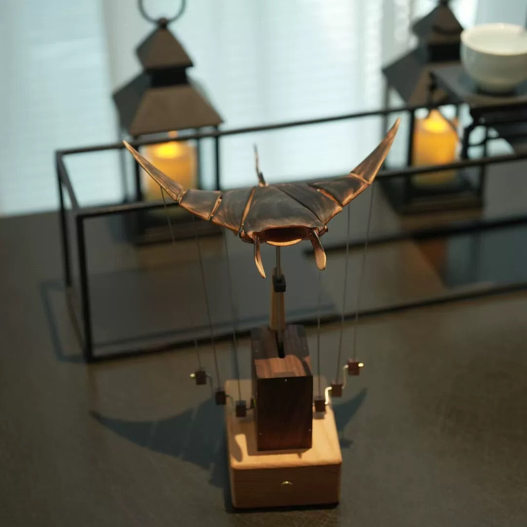 Wooden manta ray kinetic sculpture displayed on a tabletop with softly glowing lanterns in the background.